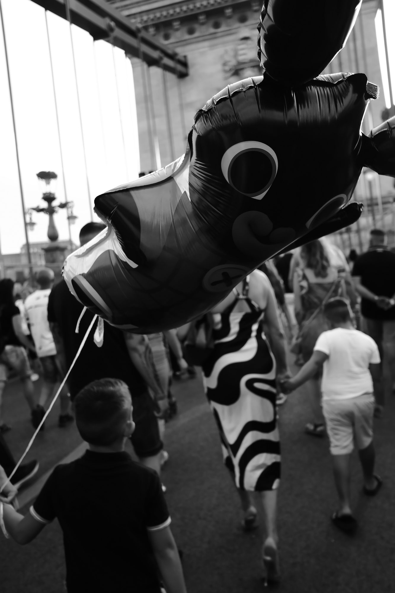 Black and white: a boy holds the string of a large cartoon-animal balloon as a crowd walks across a suspension bridge