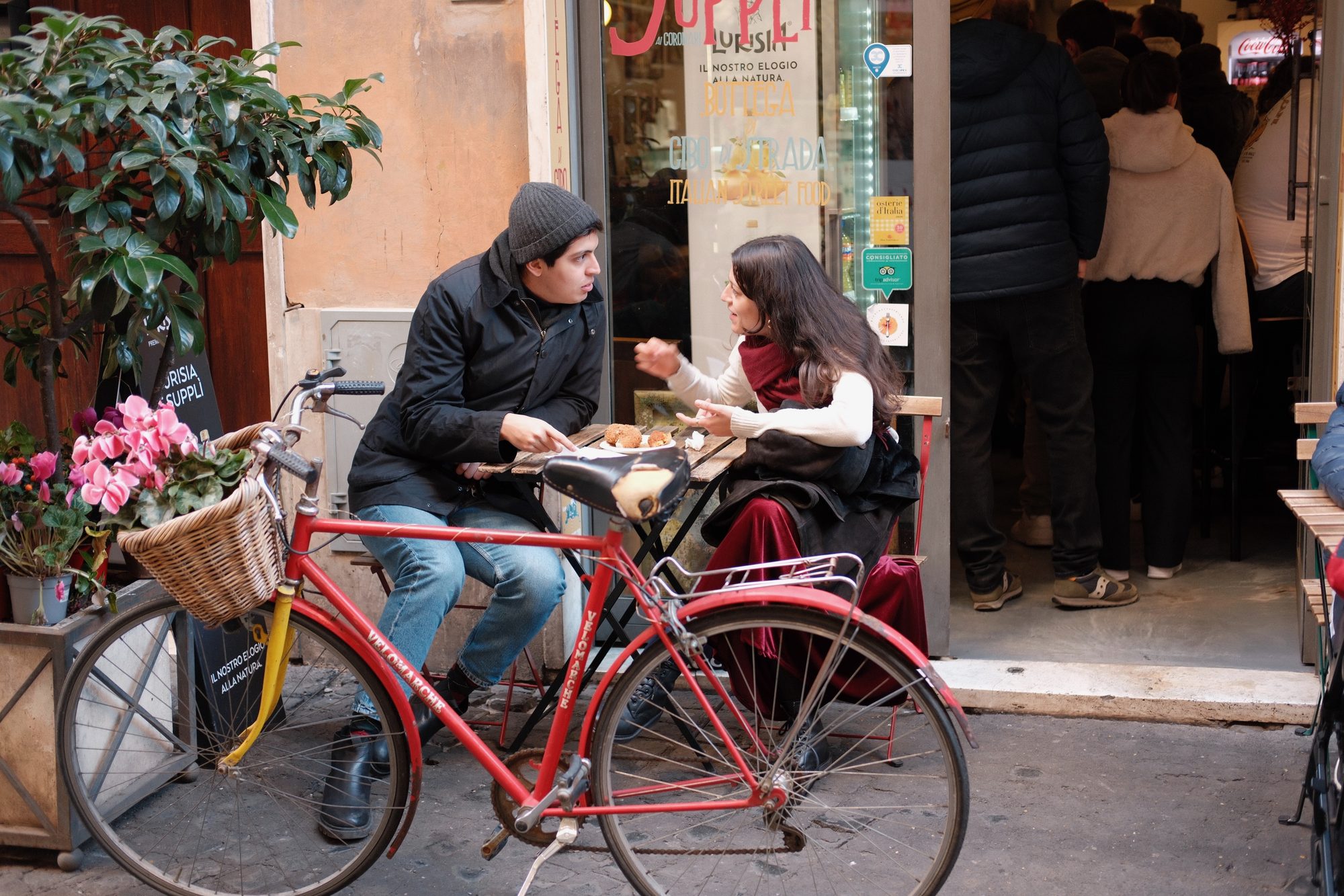 Two young people deep in conversation at a small café table outside an Italian street-food shop, a red bicycle with a wicker basket in the foreground