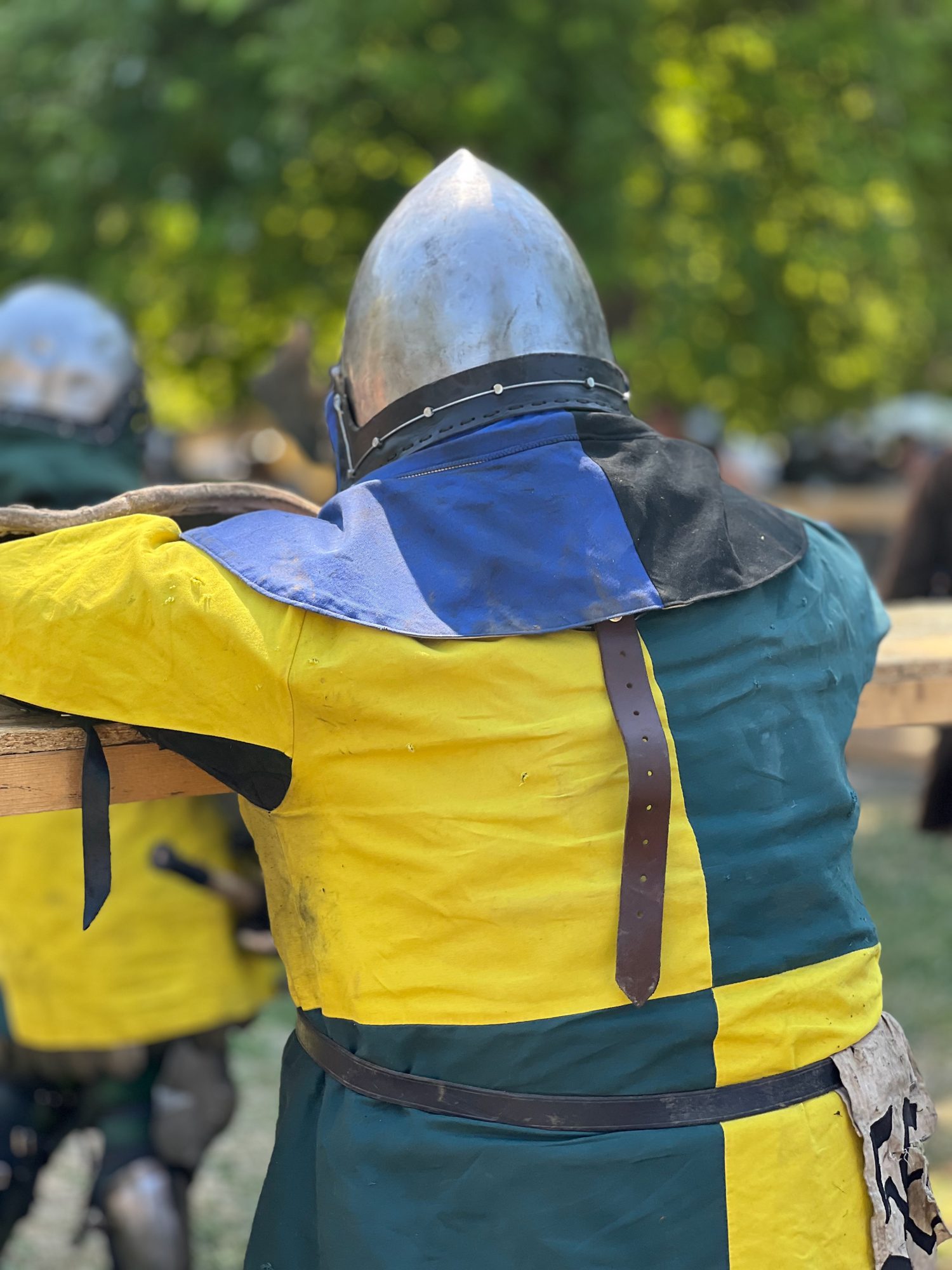 A fighter in a steel helmet and a yellow-and-green quartered tunic, seen from behind, leaning on a wooden rail at a medieval combat event