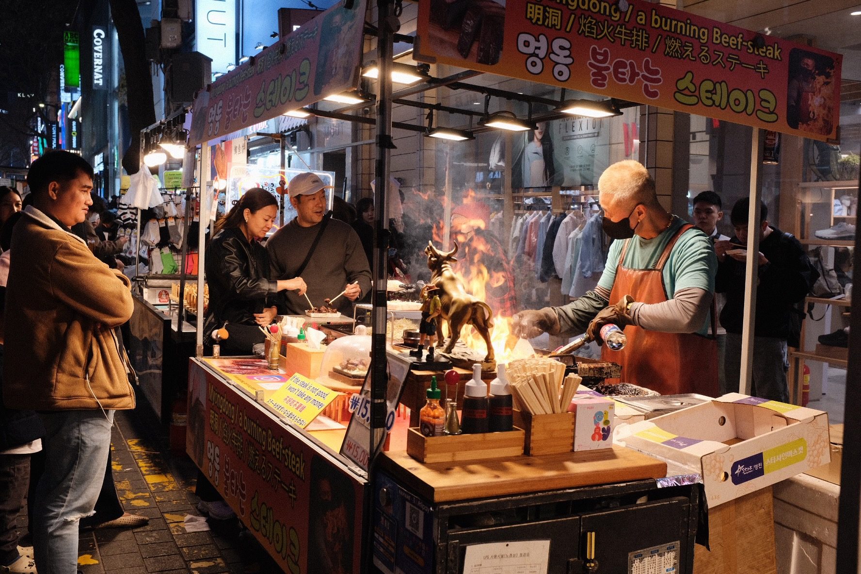 At night, a masked street vendor flames steak with a torch at a Myeongdong food stall while customers eat and watch