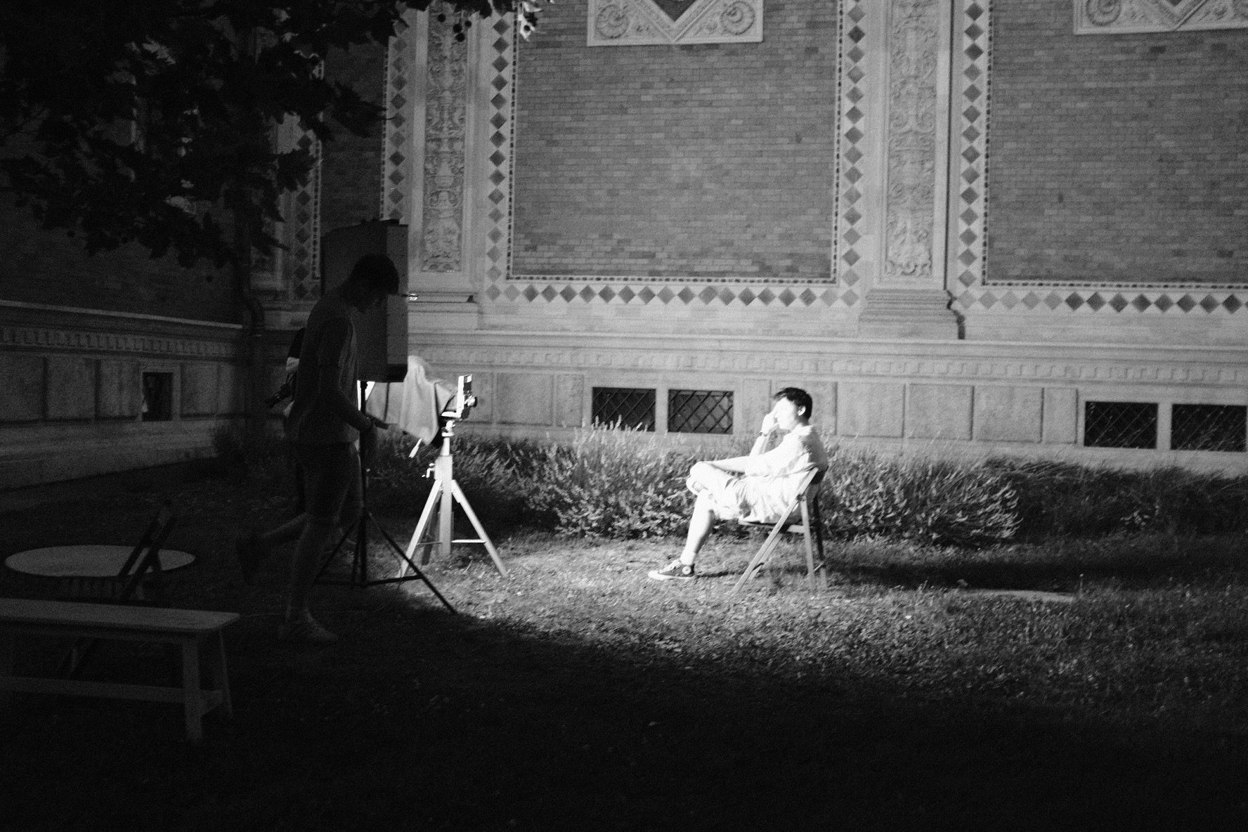 Black and white: at night, a photographer stands at a large-format camera on a tripod while a young man sits lit in a folding chair before an ornate brick facade