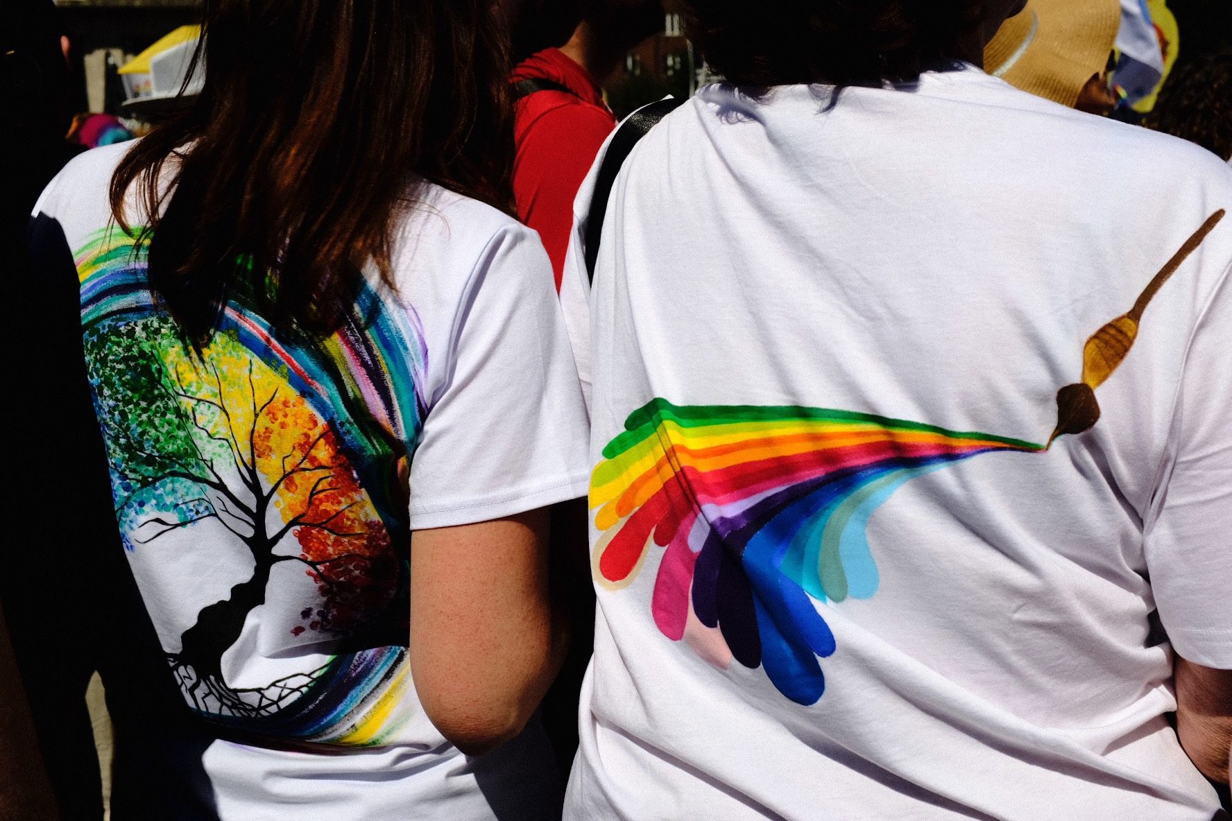Two people at a Pride event seen from behind, their white T-shirts hand-painted with a rainbow tree and a rainbow brushstroke
